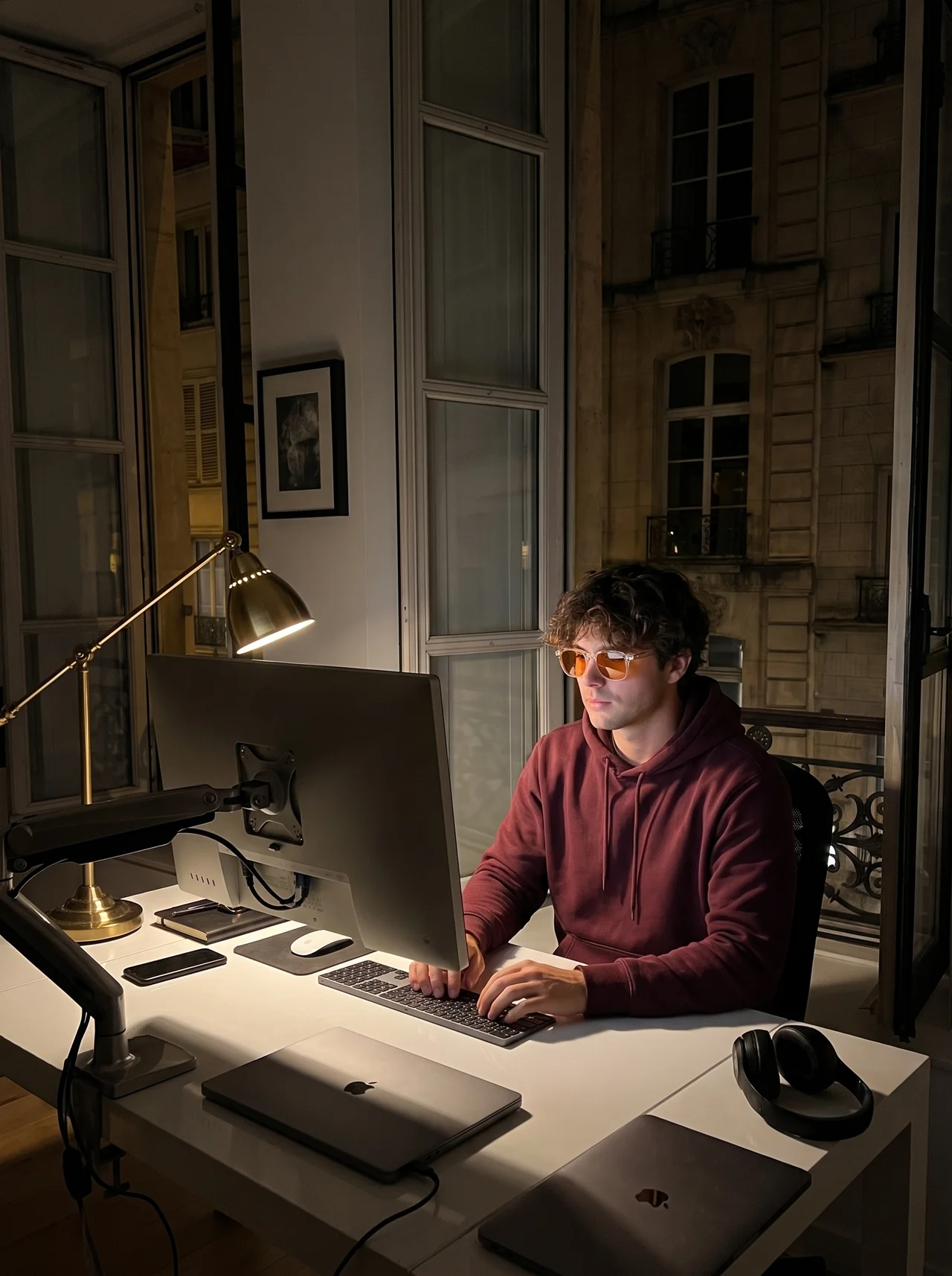 A man in amber glasses working at a lit monitor by an open window at night