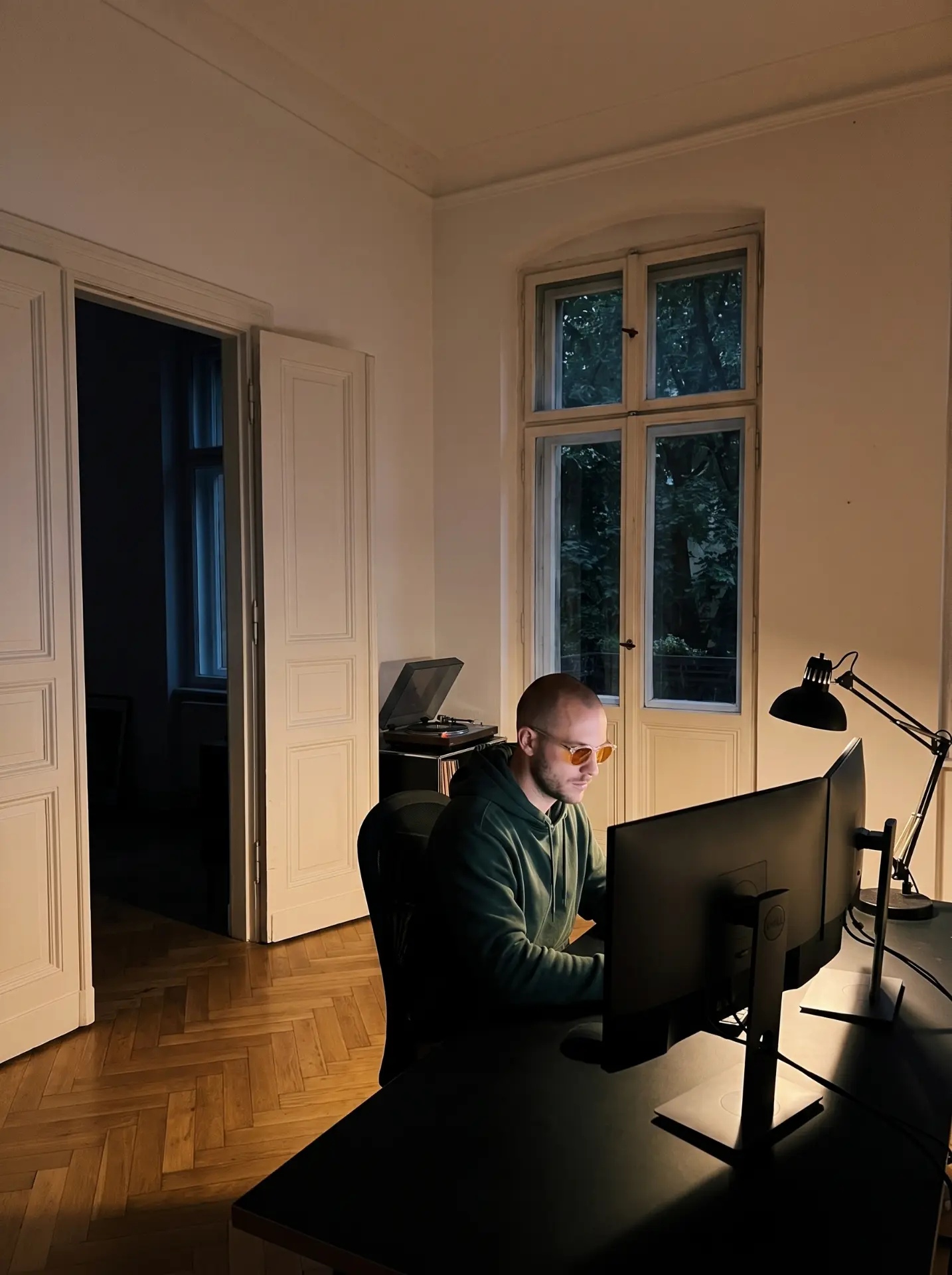 A man in amber glasses at two monitors in a quiet room with parquet floors